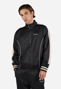 Chaqueta deportiva negra con cremallera y acentos en beige, fabricada en tejido suave. Presenta cuello alto, puños a rayas y un logo sutil en el pecho.
