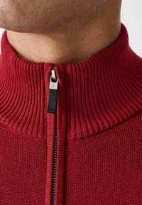 Red zip-up collar sweater with ribbed texture, featuring a silver-colored zipper and black leather pull tab. Close-up view of neckline details.