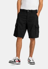 Pantalones cortos cargo negros con múltiples bolsillos, hechos de tejido de punto, con cierre de botón y un ajuste relajado. Llevados con zapatillas negras y rojas.