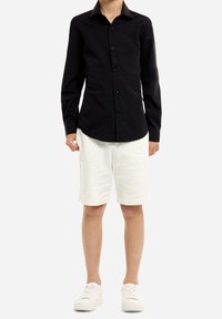 Chemise noire à manches longues avec col standard, associée à un short blanc à hauteur de genoux et des baskets blanches. Tenue simple et décontractée.