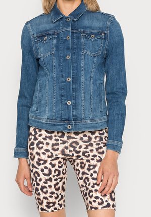 Kvinde iført blå denimjakke med metal knapper og leopardprint tætsiddende shorts står mod en ensfarvet baggrund.