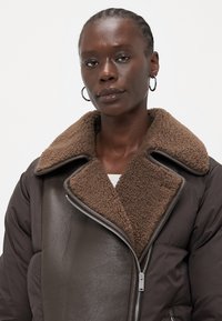 Brun skinnjakke med puffy design og brun shearling-krage. Har en sølvfarget glidelås og et lagdelt, oversized design.