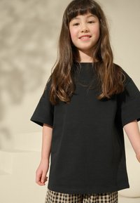 Černé tričko s krátkým rukávem, volný střih, kulatý výstřih. V páru s kostkovanými kalhotami, jemná textura, na neutrálním pozadí.