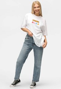 Camiseta blanca de manga corta con un gráfico colorido de "Pac-Man"; combinada con jeans rectos de mezclilla azul claro y zapatillas de lona negras.
