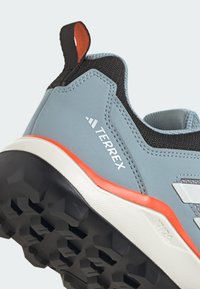 Szare buty outdoorowe z teksturowaną cholewką, czarnymi akcentami, pomarańczowymi detalami i solidną czarną podeszwą z wyraźnym bieżnikiem oraz logo.