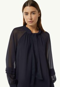Marinebluse aus leichtem Stoff mit hohem Kragen, Bindedetail und transparenten langen Ärmeln, mit gerafften Akzenten an den Schultern.