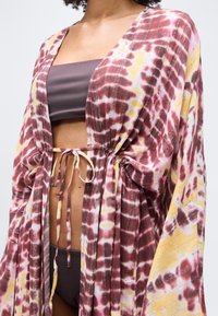 Γυναίκα που φορεί μπικίνι σε καφέ μπούστο και σλιπ, με ένα μακρύ, φαρδύ κάλυμμα με μοτίβο tie-dye σε ροζ, κίτρινο και άσπρο, δεμένο στη μέση.