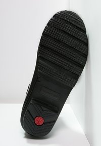 Czarna gumowa podeszwa buta z teksturowanym wzorem gripowym, poziomymi rowkami i okrągłym czerwonym akcentem z logo.