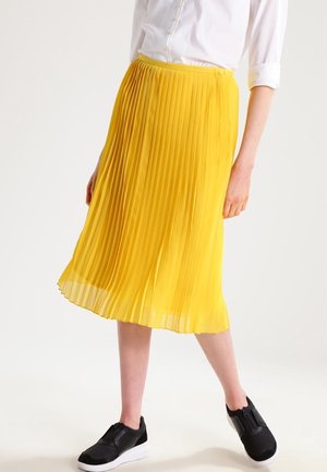 Femme portant une jupe midi plissée jaune, une chemise blanche boutonnée aux manches retroussées, et des baskets noires à enfiler avec des semelles blanches.