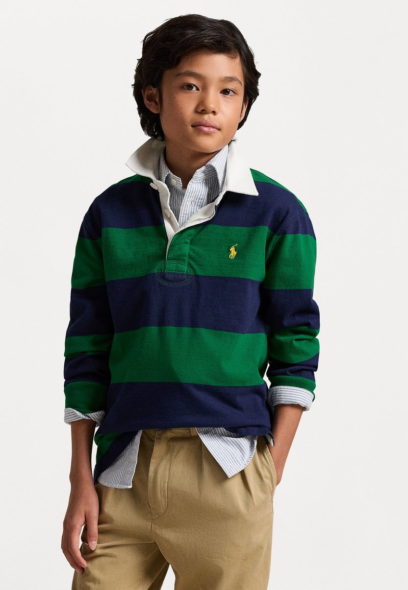 Ριγέ ράγκμπι μπλούζα σε ναυτικό μπλε, πράσινο και λευκό, με γιακά και μικρό λογότυπο. Συνδυασμένη με ανοιχτόχρωμα, πλισέ παντελόνια.