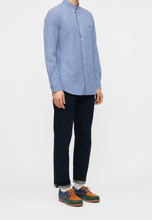 Personne portant une chemise à boutons rayée bleue et blanche, un pantalon foncé retroussé, des chaussettes blanches et des chaussures multicolores bleues, orange et vertes.