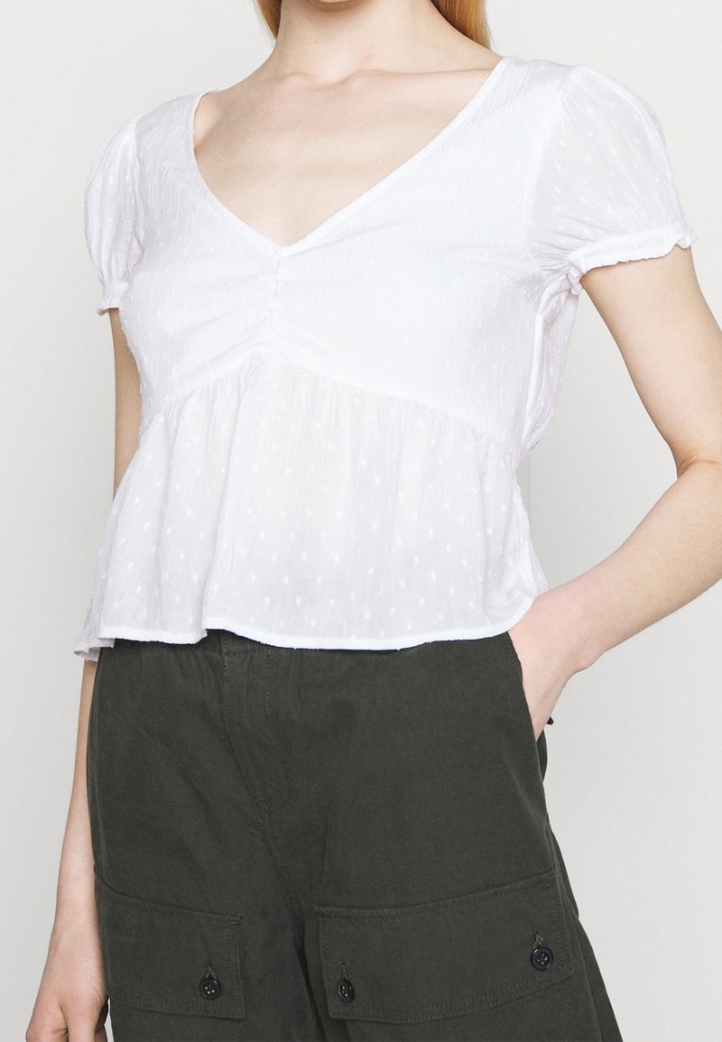 Haut blanc à manches courtes avec un col en V, tissu texturé et détail froncé au niveau de la poitrine, style péplum fluide, porté avec un pantalon vert foncé.