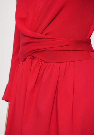 Piros ruha, átlapolt kialakítással, összehúzott derékrésszel és bő szoknyával. Simább anyag, puha tapintású, hosszú ujjakkal.