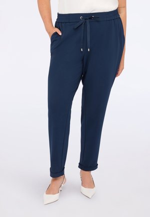 Frau in marineblauen Jogginghosen mit hochgerollten Säumen und weißen spitzen Slingback-Schuhen, steht mit einer Hand in der Tasche.