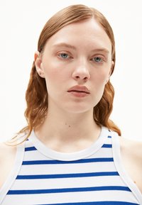 Witte tanktop met blauwe horizontale strepen, geribde textuur, ronde halslijn en geen zichtbare bevestigingen. Model heeft schouderlang golvend haar.