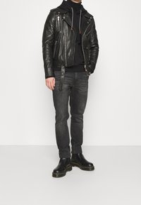 Giacca da biker in pelle nera con cerniere argentate, sovrapposta a una felpa nera con cappuccio, abbinata a jeans grigio scuro e stivali Chelsea neri.