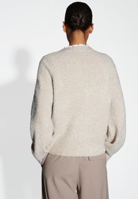 Persoon met een knot in het haar, gekleed in een lichtbeige gebreide trui en taupe broek, staand met de handen in de zakken, weg van de camera.