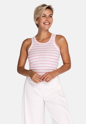 Smiling woman with short blonde hair wearing a sleeveless pink and white striped top and high-waisted white pants, posing against white background.