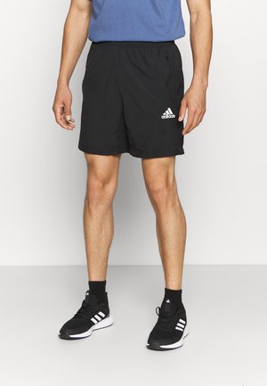 Pantaloni corti sportivi neri con vita elastica, dotati di tasche con zip e logo Adidas bianco. Indossati con sneakers nere e calze.