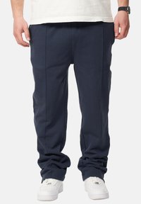 Marineblaue, lässig geschnittene Hose aus weichem Material mit glatter Textur, mit vorderen Falten und elastischem Bund. Getragen mit weißen Sneakers.