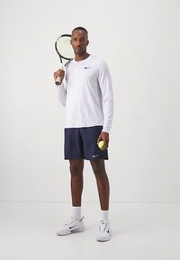 Biely športový top s dlhým rukávom, tmavomodré šortky s logom Nike, držiaci tenisovú raketu a žltý tenisový loptičku, obutý v bielych teniskách.