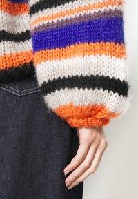 Pull en maille avec larges manches rayées orange, violet, blanc et noir. Présente une texture duveteuse et des poignets côtelés. Tissu en denim foncé visible.