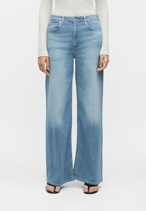 Φαρδιά τζιν ανοιχτού μπλε χρώματος, φορεμένα με λευκό μακρυμάνικο τοπ και μαύρα σανδάλια ανοιχτής μύτης, εμφανιζόμενα από τη μέση και κάτω.