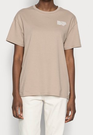 Person iført en løs beige t-shirt med lille hvid tekst øverst til venstre og hvide bukser, stående mod en ensfarvet lys baggrund.