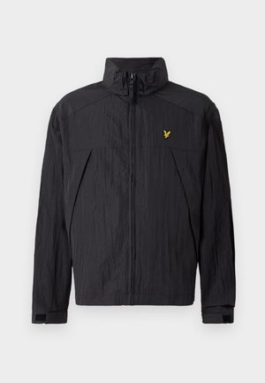 Veste zippée noire avec une texture, dotée d'un col montant et d'un petit logo doré sur le côté supérieur gauche.
