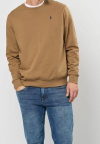 Sudadera marrón con un cuello redondo acanalado y puños, que presenta un pequeño logo azul marino en el pecho. Combinada con jeans de mezclilla azul.