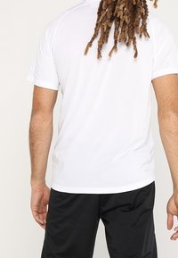 Camiseta deportiva blanca de manga corta con una textura suave; presenta un escote redondo y un corte relajado, combinada con pantalones cortos negros.