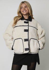 Cremefarvet shearlingjakke med en sort læderkrave, sorte knaplukninger og kontrasterende kanter. To store klaplommer inkluderet.