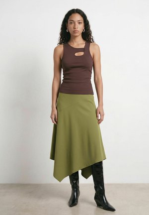 Femme aux cheveux bouclés portant un haut marron sans manches avec une découpe, une jupe asymétrique olive et des bottes noires montant jusqu’aux genoux, debout contre un mur blanc.