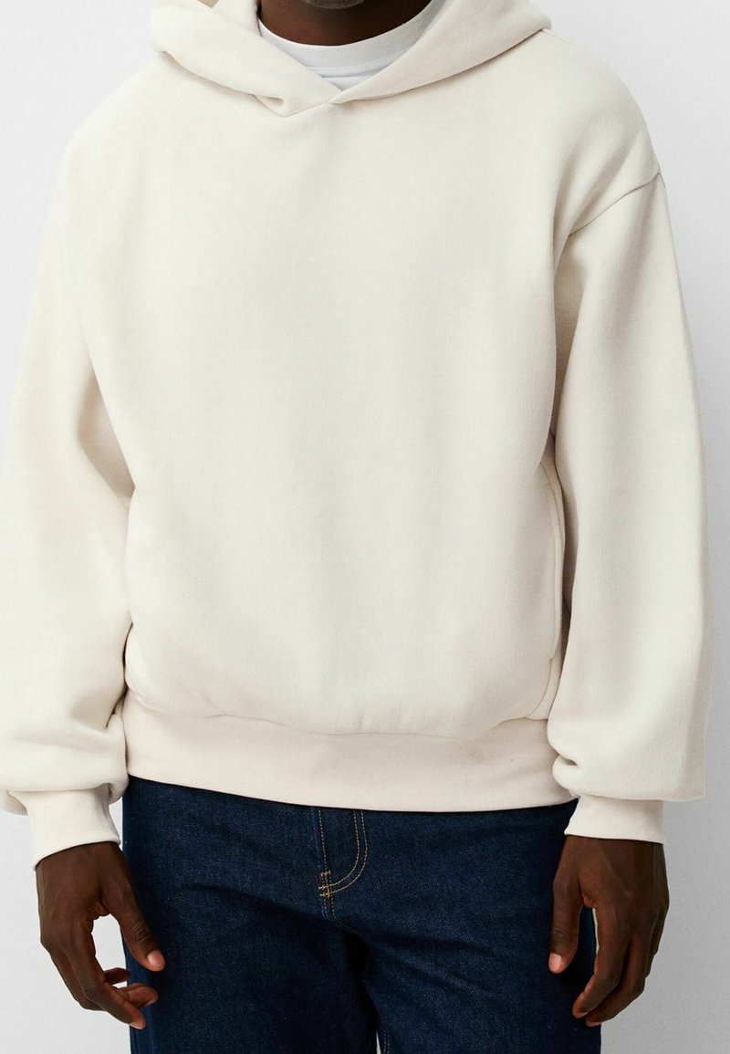 Hombre vestido con una sudadera de gran tamaño color blanco roto y jeans azul oscuro, de pie frente a un fondo sencillo.