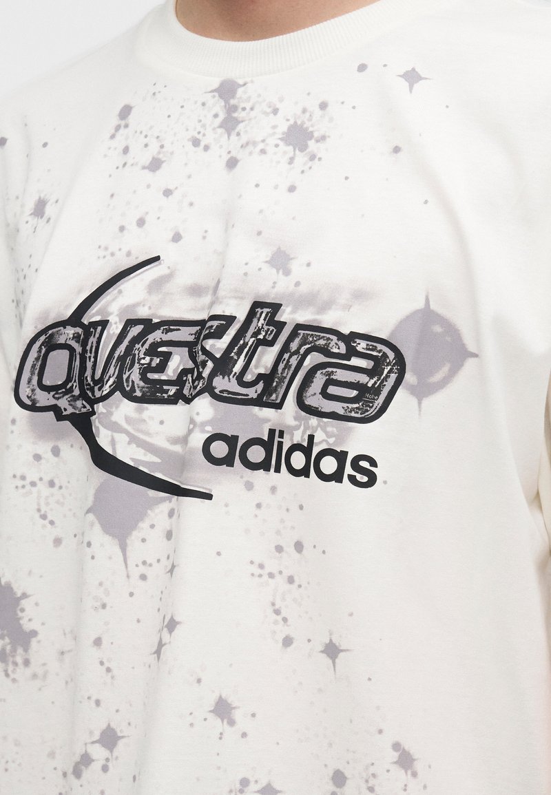 Maglietta bianca Adidas con la scritta "questra" in carattere nero stilizzato e motivi di schizzi grigi intorno al testo.