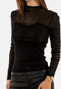 Top negro de manga larga con una tela etérea y texturizada y un escote de cuentas en forma de ondas. Combinado con pantalones negros ajustados.