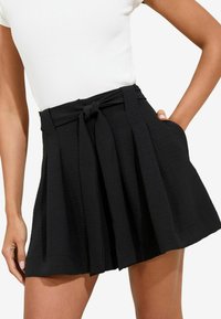 Una persona indossa un top bianco aderente e una gonna nera plissettata con una vita legata, con le mani nelle tasche, mostrando uno stile casual.