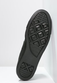 Semelle de sneaker en caoutchouc noir avec une surface texturée, présentant un motif géométrique et un logo estampé sur le côté, inclinée contre un fond blanc.