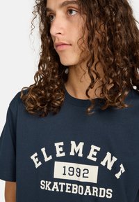 Námořnická modrá bavlněná tričko s bílou grafikou s textem "ELEMENT", "1992" a "SKATEBOARDS". Krátké rukávy, kulatý výstřih.