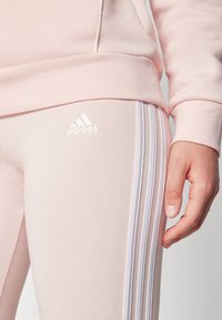 Svetlo roza atletske legice z belim tridelnim dizajnom ob strani in Adidas logotipom v beli barvi na boku. Gladka tekstura.