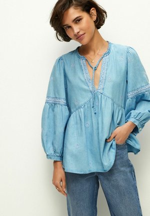 Blouse en coton bleu clair avec des manches bouffantes, motifs floraux brodés, décolleté en V avec un détail à nouer, et une coupe ample. Associée à un jean en denim.