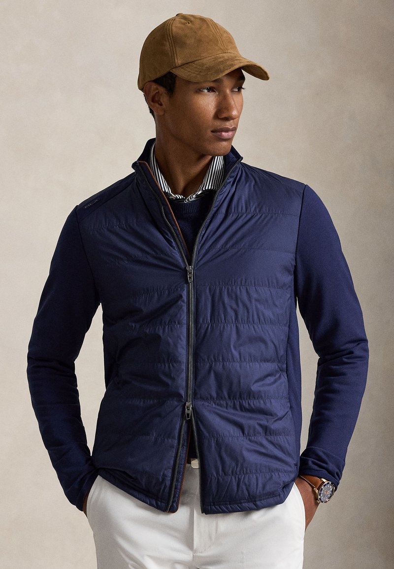 Muž nosiaci hnedú čiapku, námornícky modrú prešívanú bundu na zips cez pruhovanú golierovú košeľu, biele nohavice a náramkové hodinky, ruky v vreckách, pozerá sa doprava.