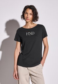 T-shirt noir à manches courtes, col rond, avec le texte blanc "FIND" sur le devant. Tissu doux, drapés subtils. Porté avec un pantalon clair.