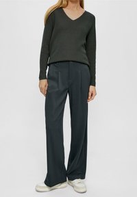 Eine Frau trägt einen dunkelgrünen V-Ausschnitt-Pullover, lockere dunkelgraue Hosen und weiße Sneakers und steht vor einem schlichten hellen Hintergrund.