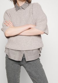 Un pull en tricot texturé gris clair à manches courtes, porté par-dessus une chemise à col rayée marron et blanche, associé à un jean gris.