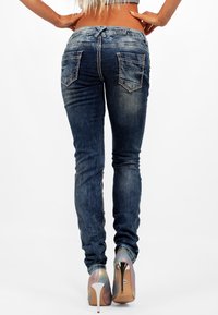 Jeans ajustados en denim azul oscuro con dos bolsillos traseros y costuras en contraste, combinados con tacones altos plateados brillantes que presentan un efecto degradado.