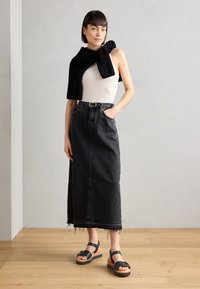 Black denim midi skirt with frayed hem, paired with a fitted white tank top. Black sweater draped over shoulders. Chunky black sandals.
