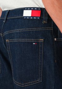 Tamno plave traperice s žutim šavovima, s logom Tommy Jeans na pojasu i malom crveno-bijelo-plavom oznakom na stražnjem džepu.