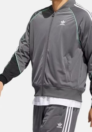 Person trägt einen grauen Adidas-Trainingsanzug mit weißen Streifen an Ärmeln und Hosen sowie hellblauen Paspeln an der Jacke.