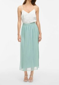 Bílá saténová košilka s tenkými ramínky, spárovaná s volnou maxi sukni v mátově zelené barvě vyrobenou z jemné, lehké látky. Stříbrné sandály na pásky.